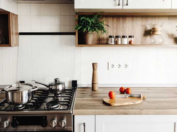 Kitchen worktop with stove and pans on top, cutting board with tomatoes and a knife, shelves with spices and plants